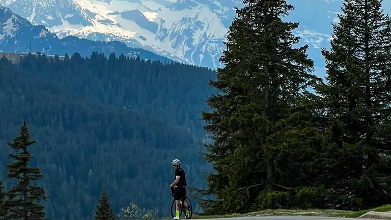 En cyklist kigger sig over skulderen. De står alene og står stille. De står ved et stort træ. Der er bjerge i baggrunden.
